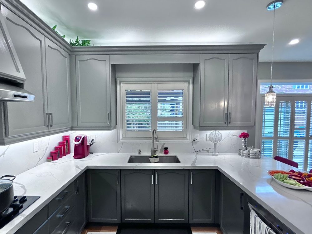 A kitchen sink under a window surrounded by grey cabinets and white quartz
