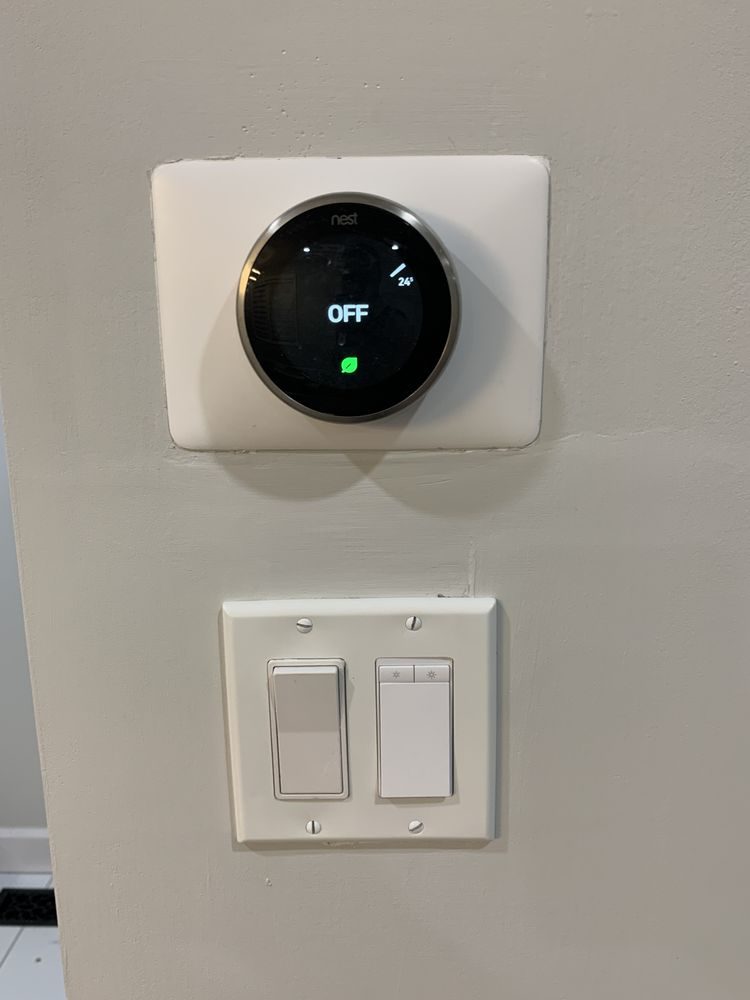 A smart thermostat above a double light switch on a grey wall